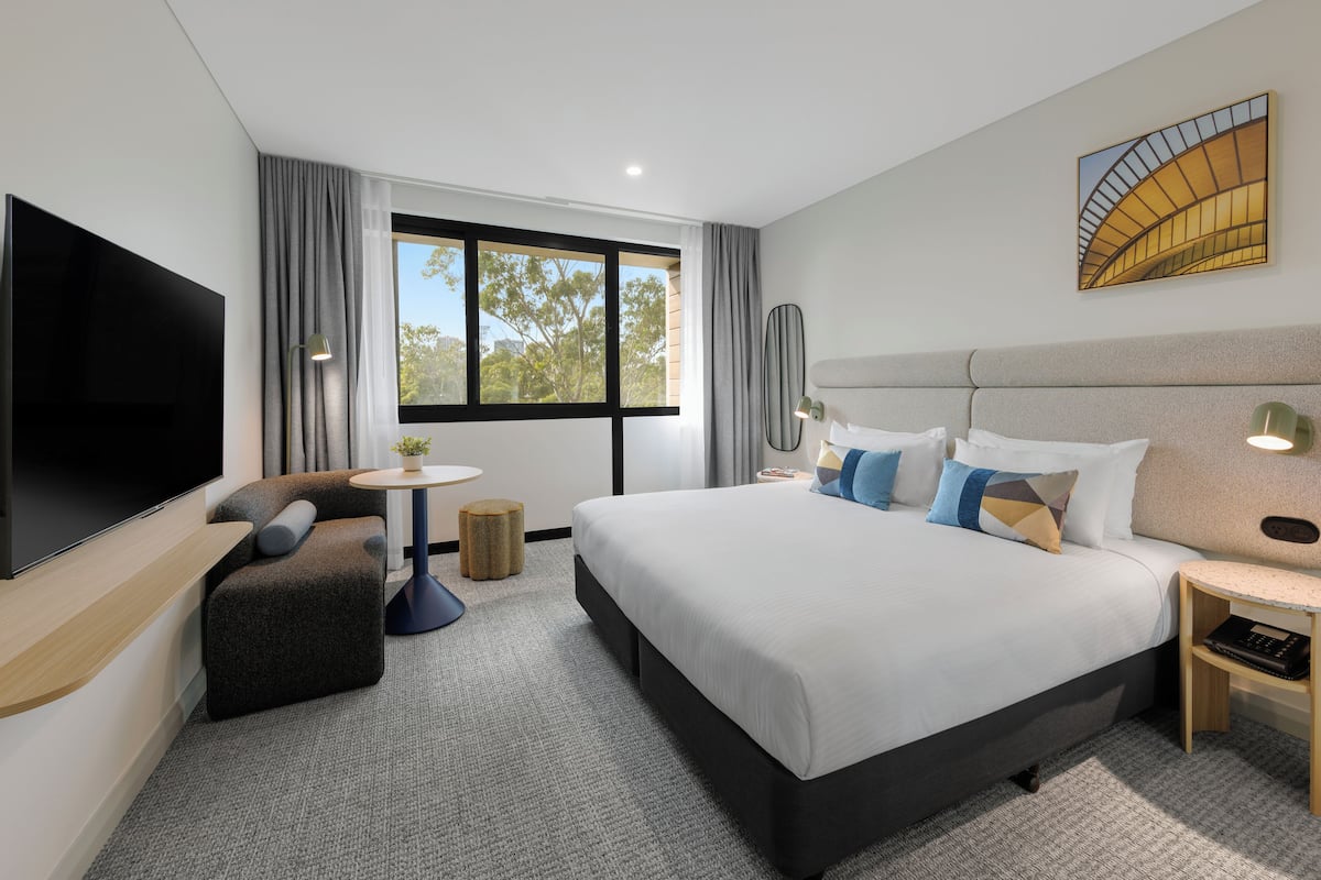 A modern hotel room with a large bed, a flat-screen TV, a small round table, a sofa, and a large window.