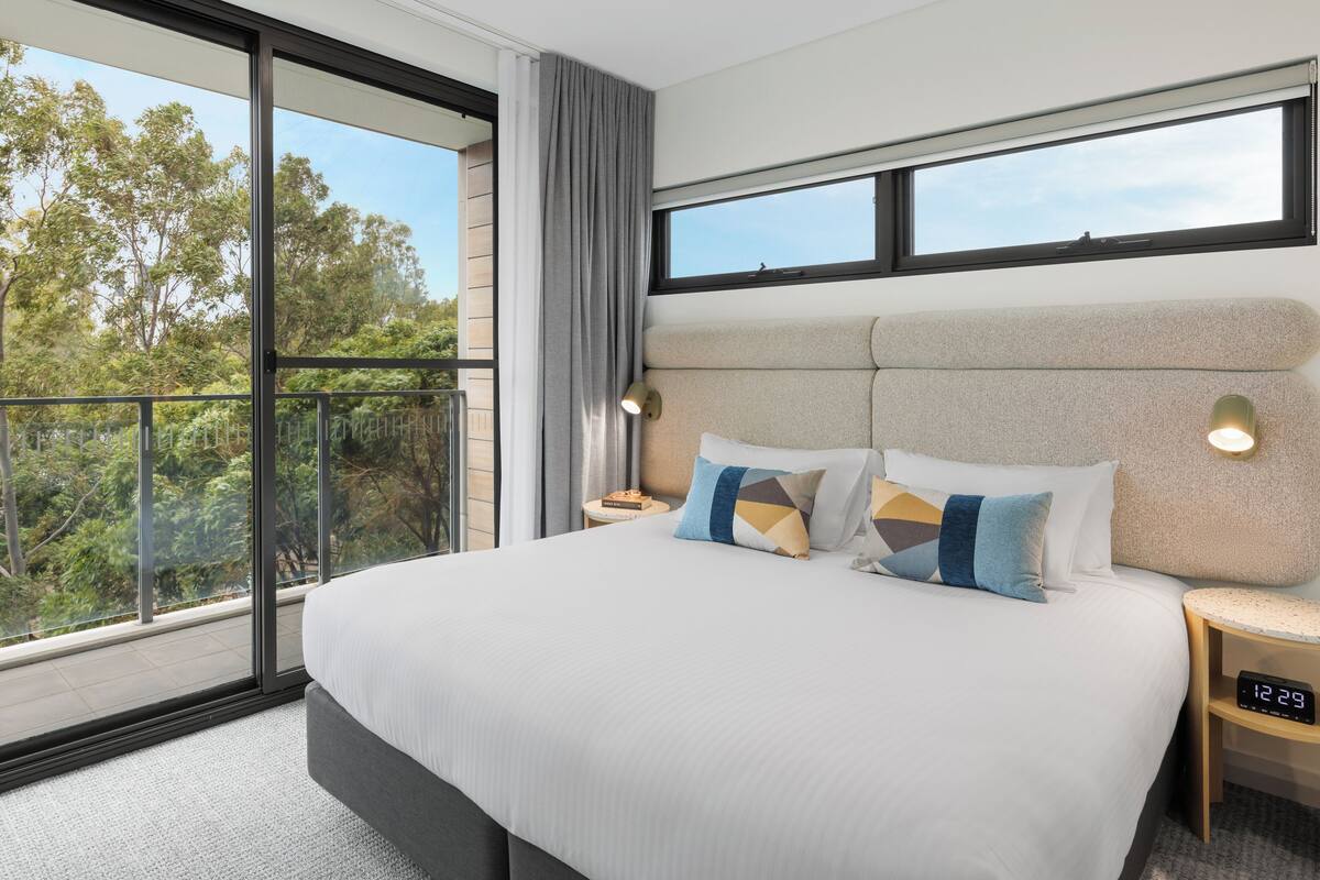 A modern hotel room with a large bed, two bedside tables, a digital clock, and a balcony with a view of trees.