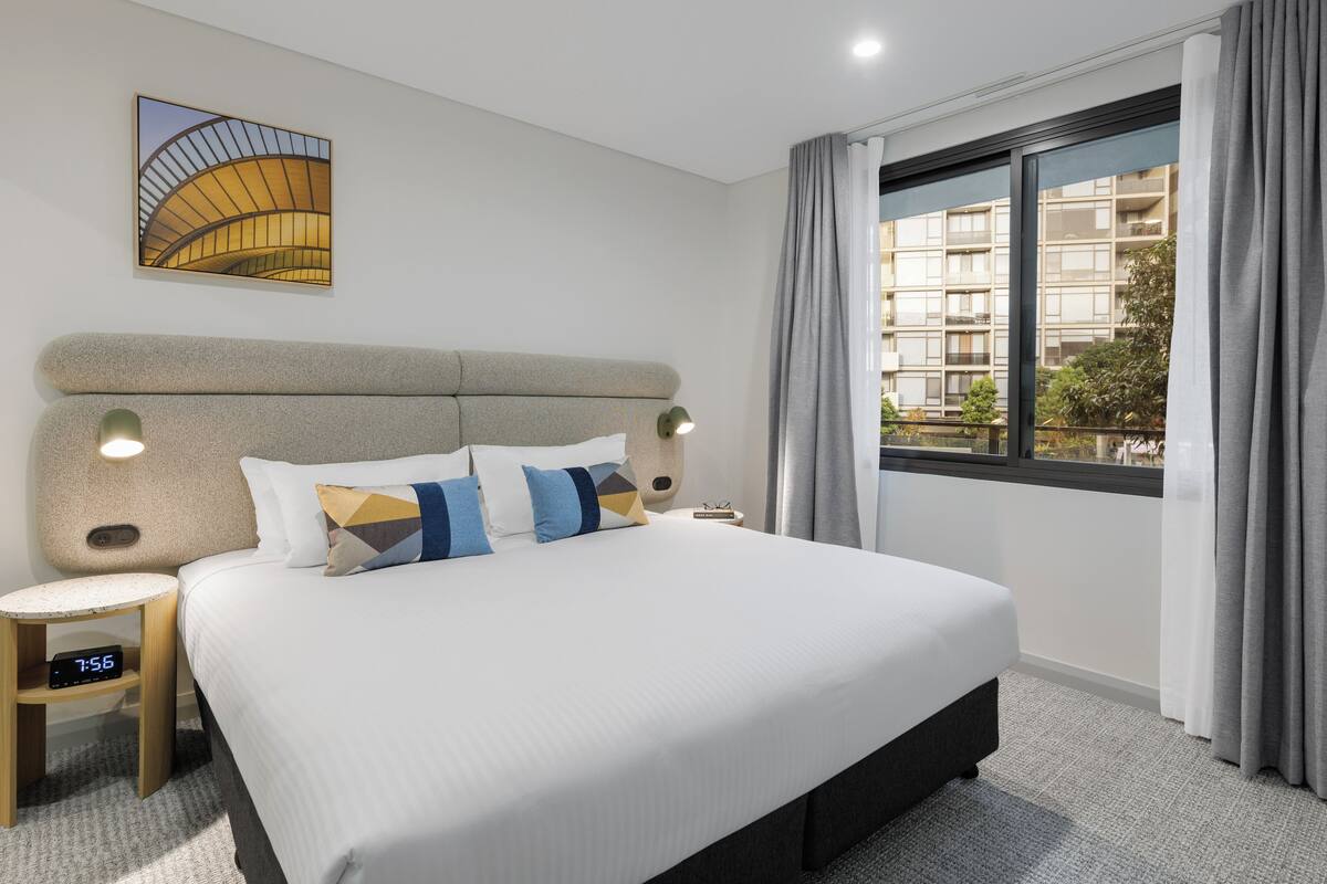 A hotel room with a large bed, a nightstand, a clock, a window with curtains, and a wall art piece.