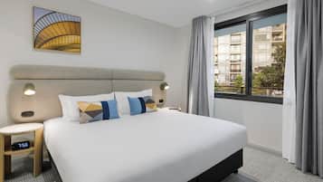 A hotel room with a large bed, a nightstand, a clock, a window with curtains, and a wall art piece.