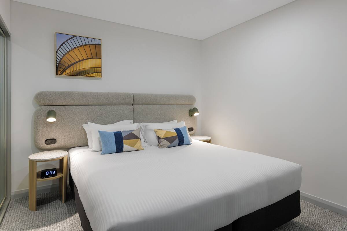 A modern hotel room with a large bed, two bedside tables, a clock, and a wall-mounted artwork.