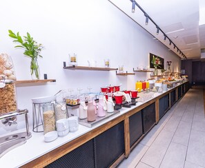 Tarjolla on monipuolinen buffet, jossa on erilaisia ruokia ja juomia, sekä koristeeksi asetettu kasvi ja kahvipiste.