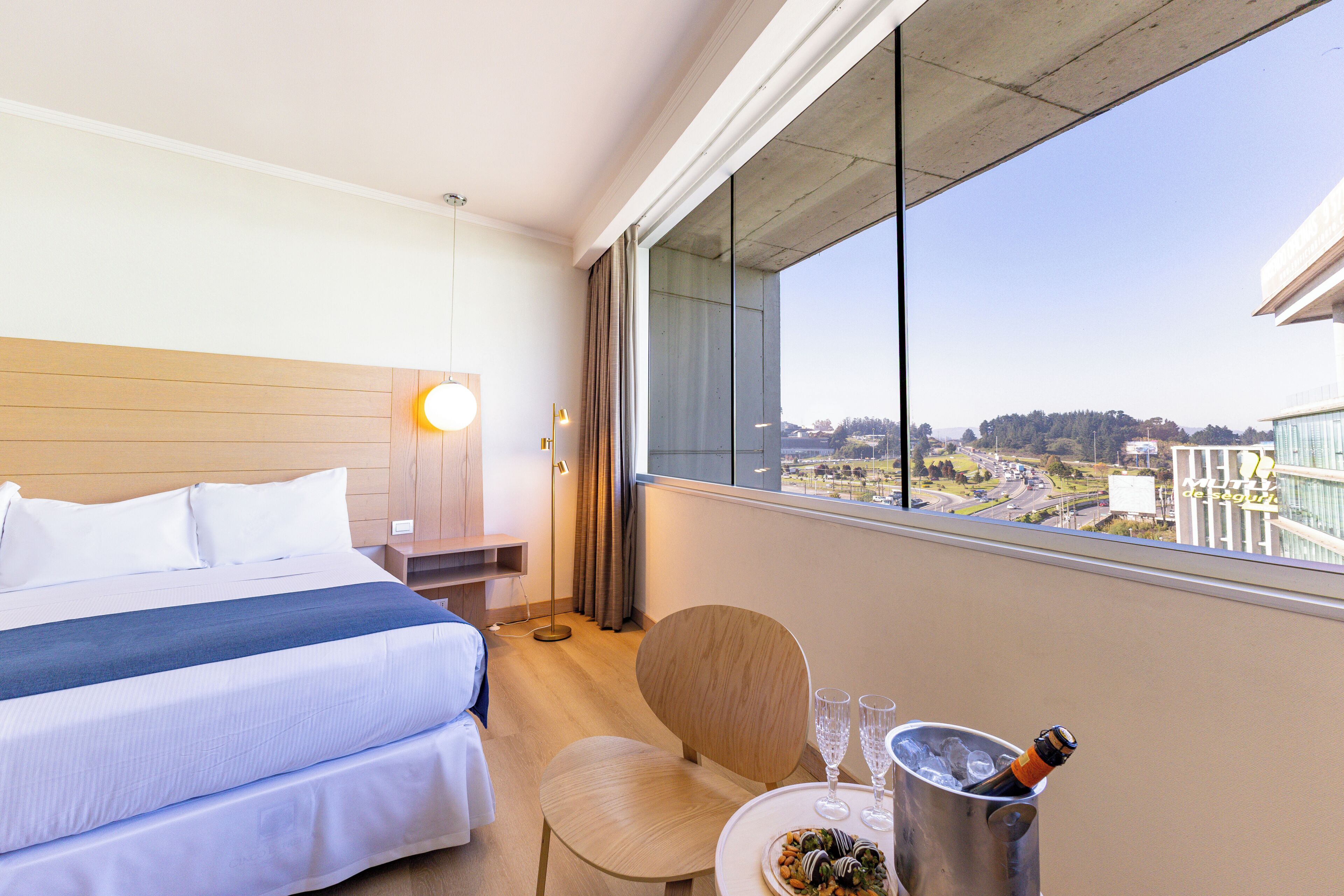 A hotel room with a bed, bedside table, lamp, chair, and a view of the city through a large window.