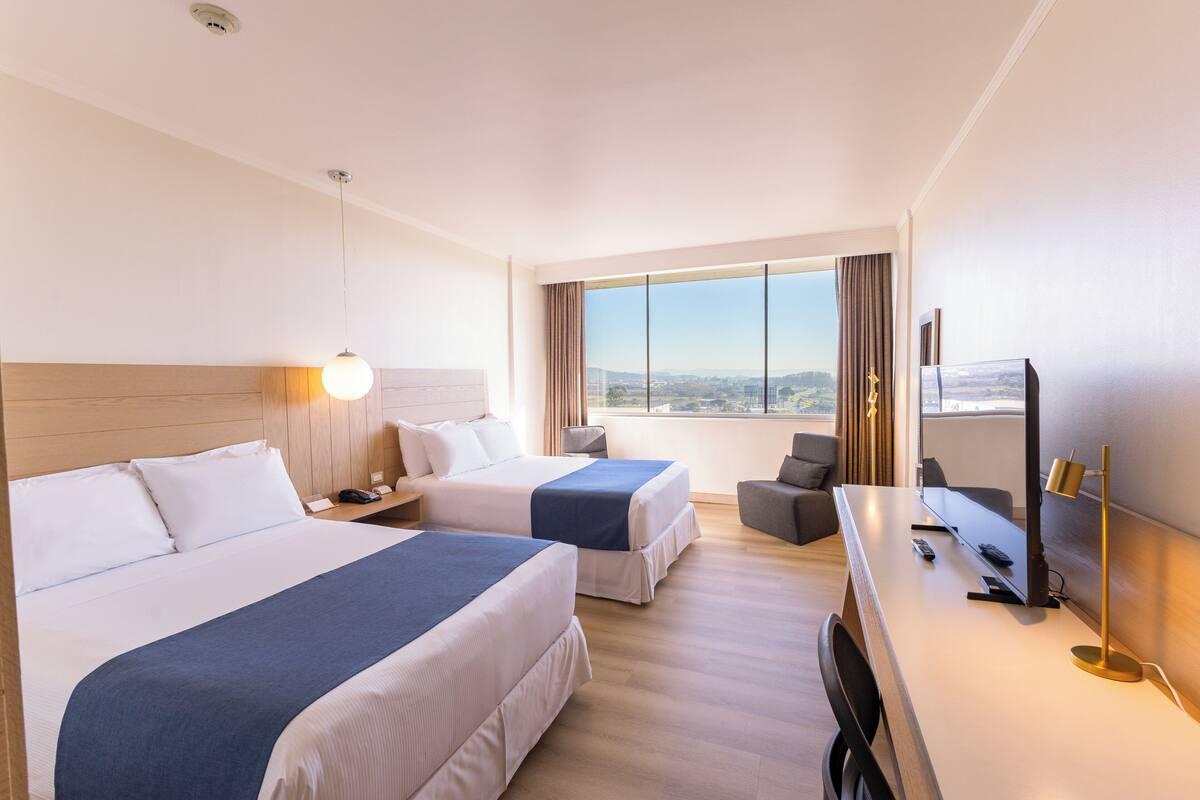 A hotel room with two beds, a desk with a TV, and a large window with a city view.