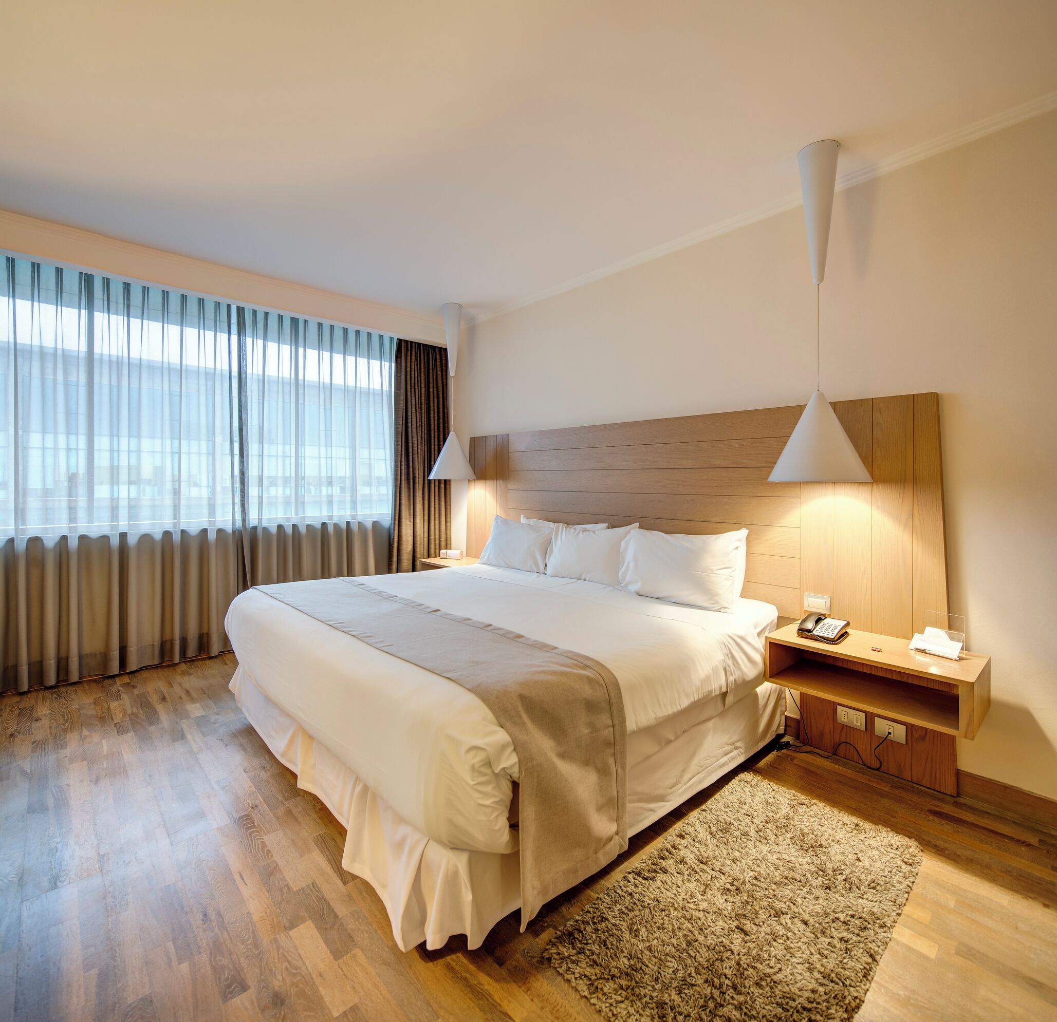 A hotel room with a large bed, a bedside table with a lamp, a telephone, and a small desk with a notepad and pen.