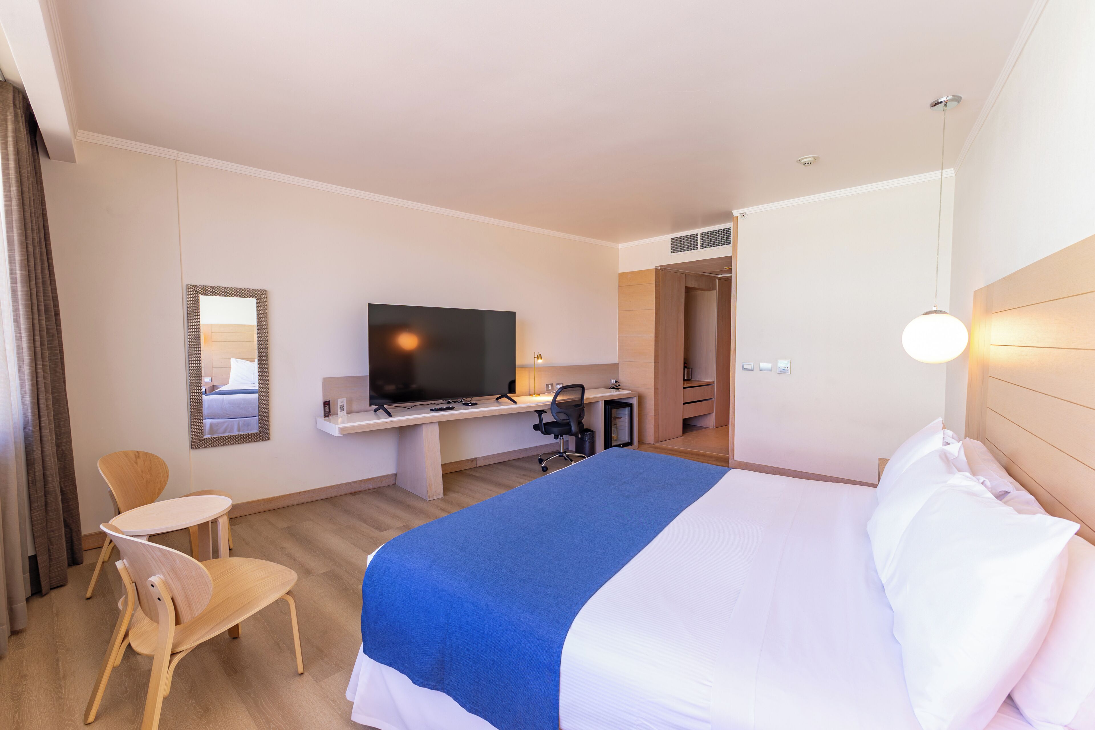 Una habitación de hotel con una cama, un escritorio con televisor, una silla y un espejo.