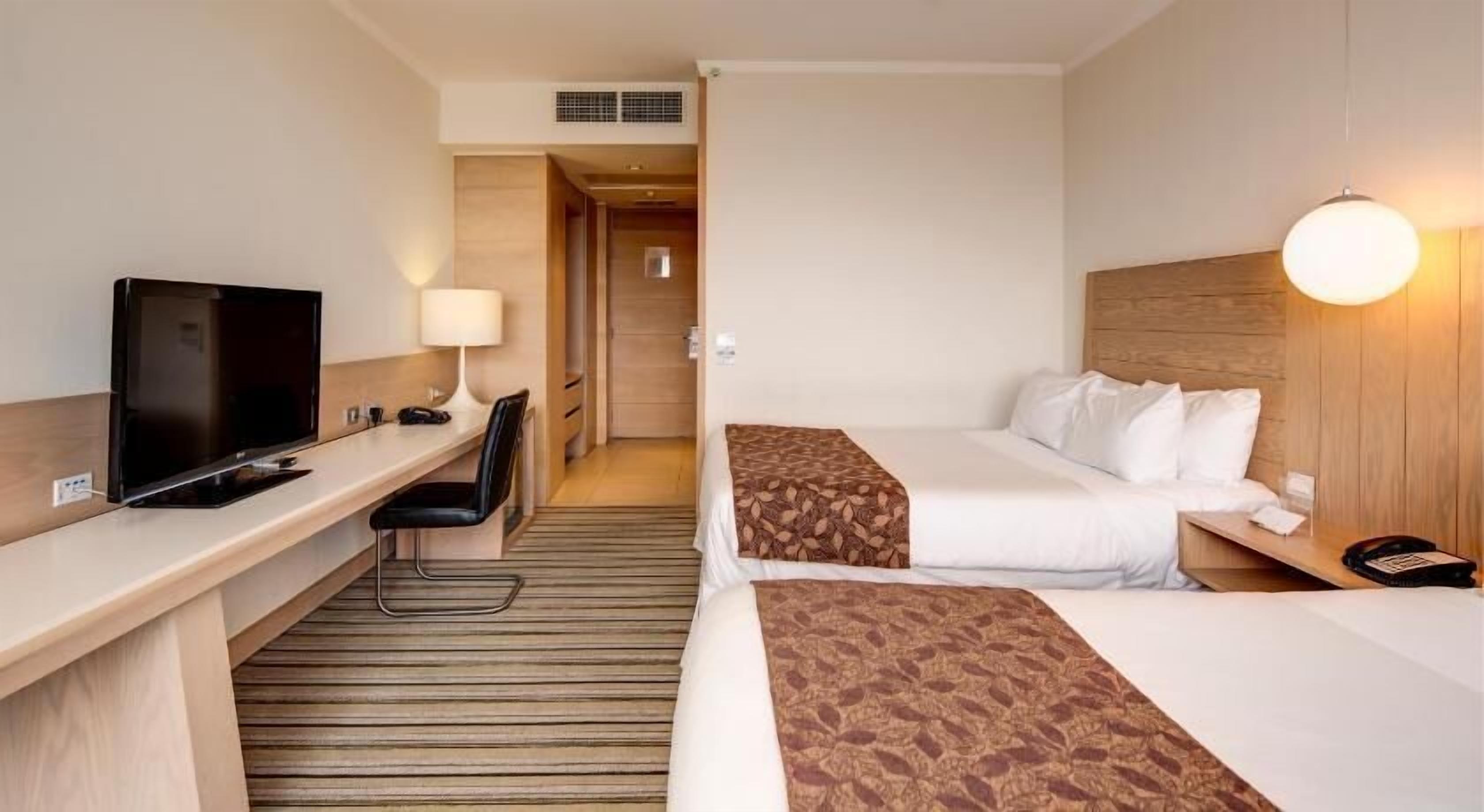 A hotel room with a desk, TV, two beds, a chair, and a lamp.