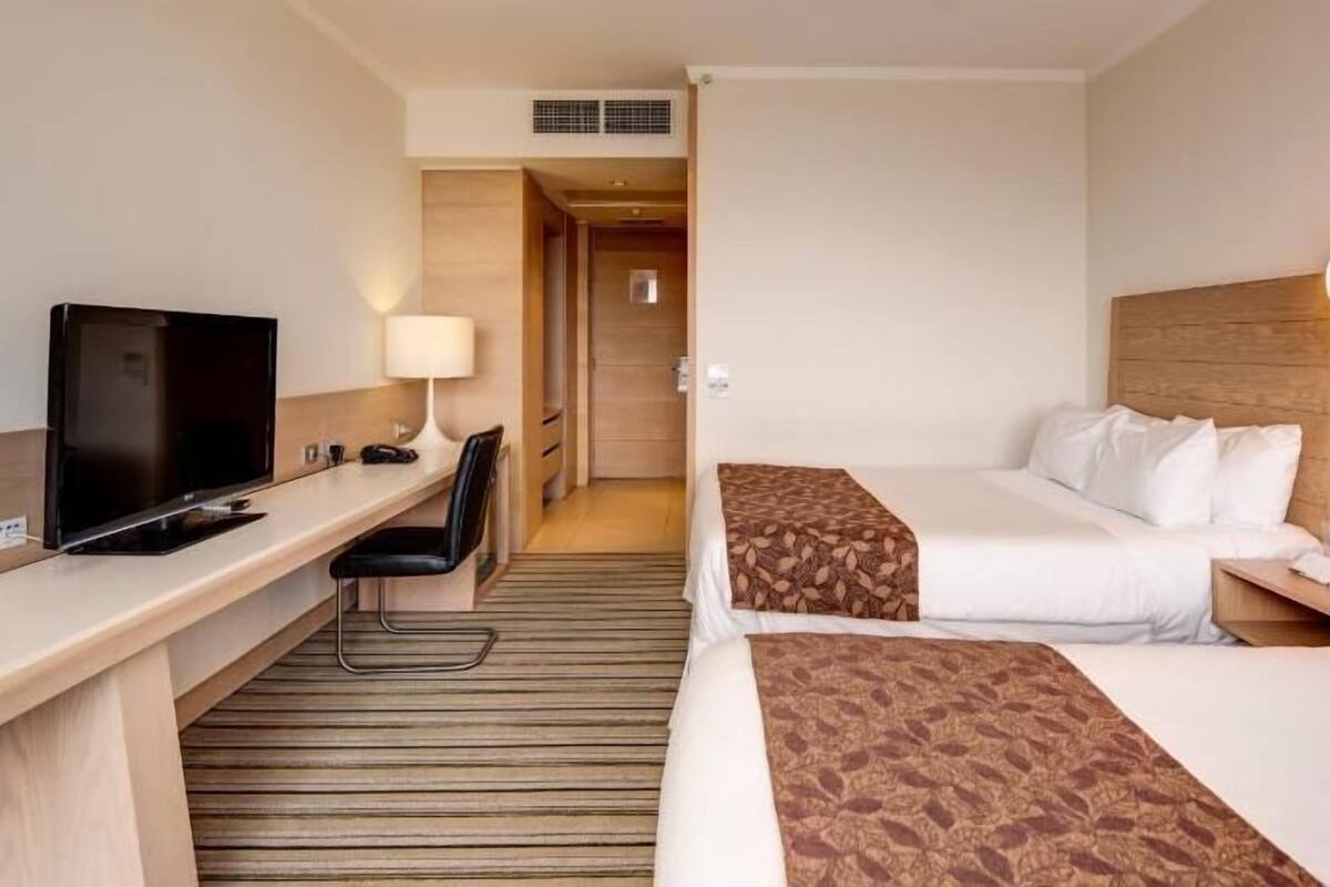 A hotel room with a desk, TV, two beds, a chair, and a lamp.