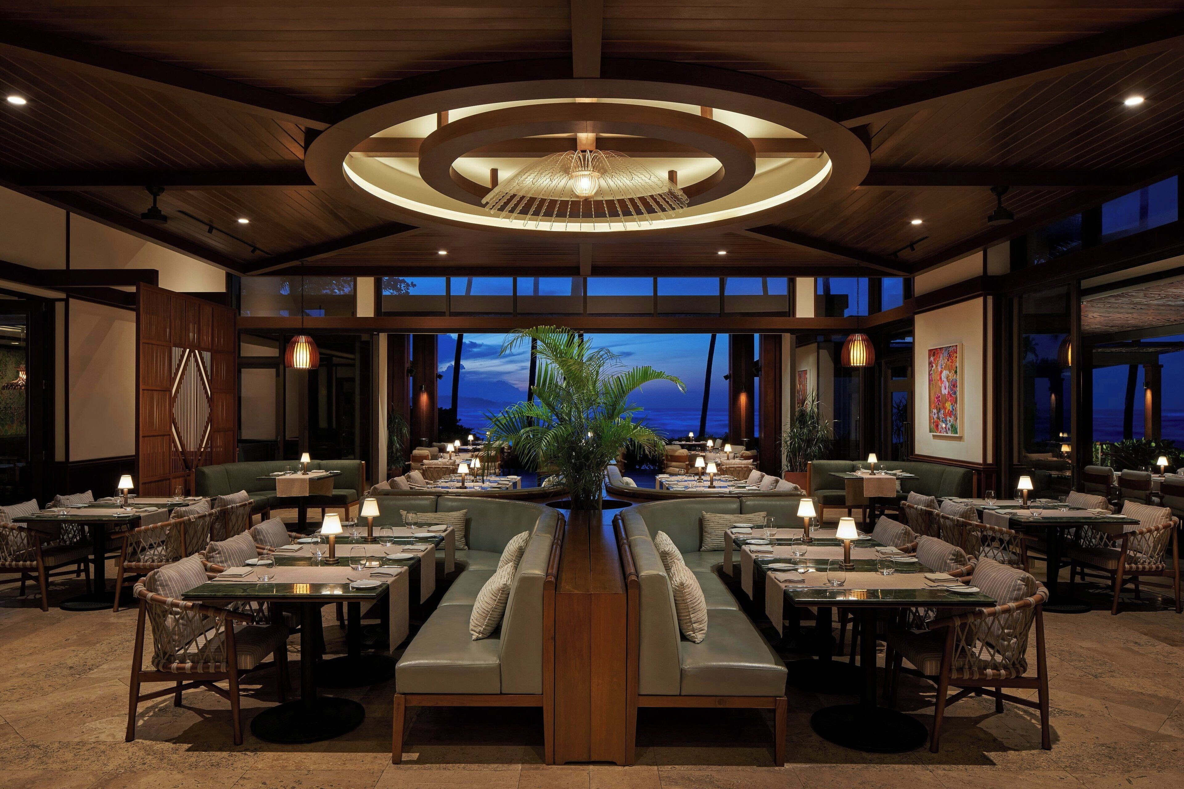 A spacious restaurant with a central seating area, multiple tables, and a large view of the ocean at dusk.