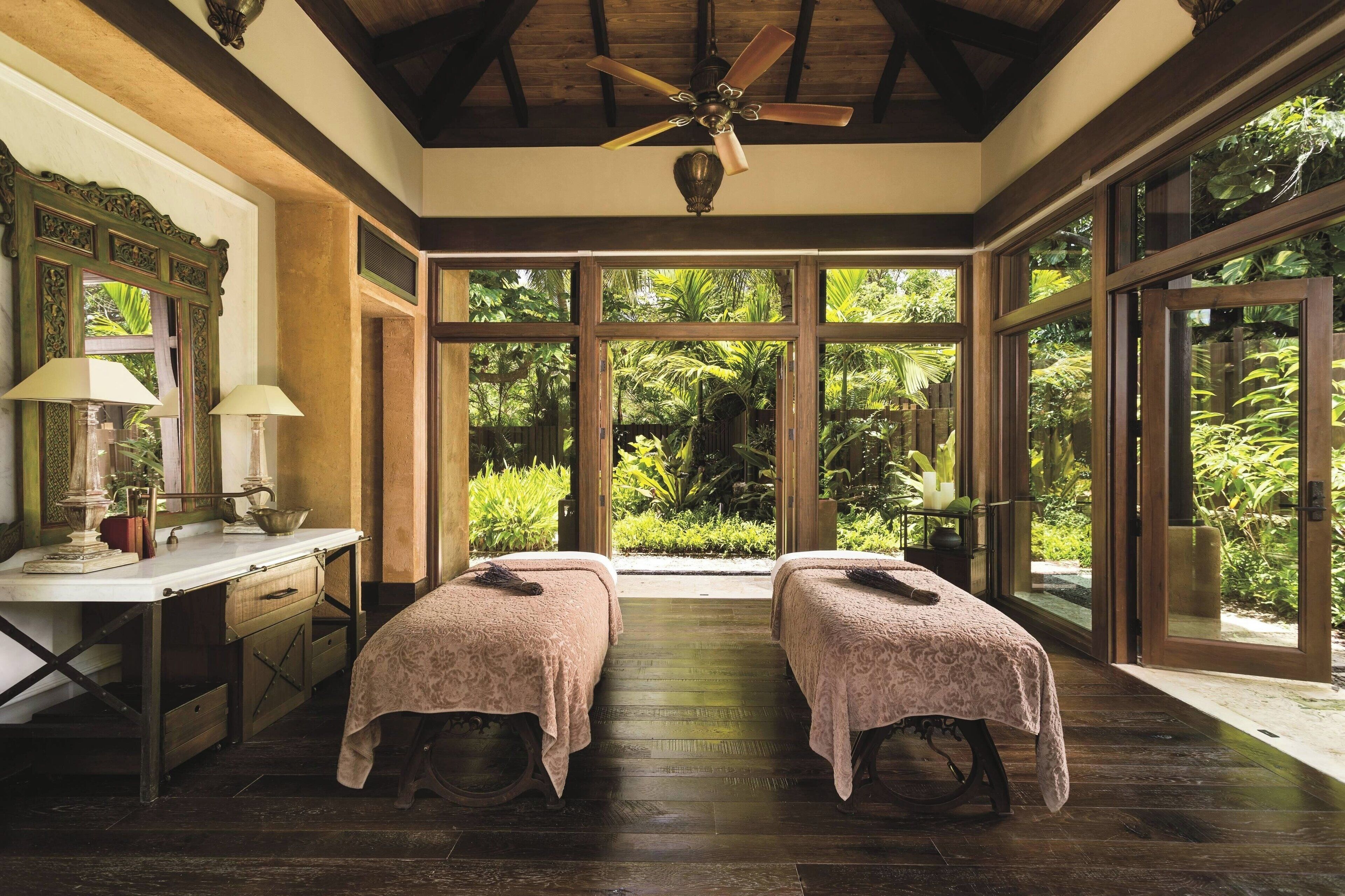 A room with two beds, a desk, and large windows overlooking a garden.
