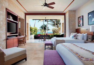 A hotel room with a bed, a desk, a TV, and a balcony with a view of palm trees and the ocean.