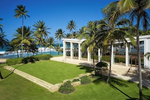 A modern beachfront property with a well-maintained lawn, palm trees, and a glass-enclosed building with a staircase leading to the entrance.