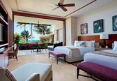 A hotel room with two beds, a ceiling fan, a balcony with a view of the ocean, and artwork on the walls.
