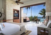 A bedroom with a bed, a TV, a sofa, and a balcony with a view of the ocean and palm trees.