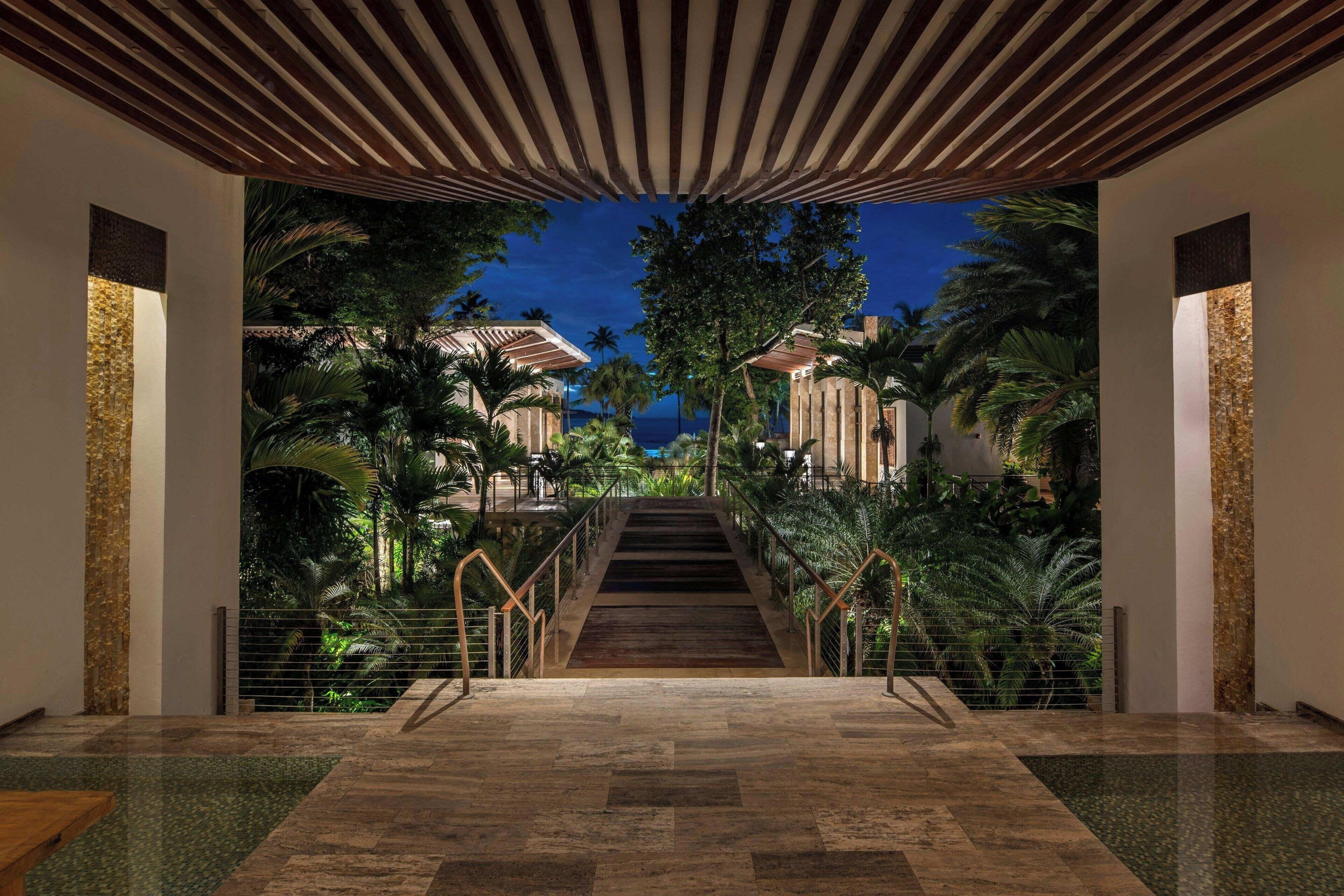 A modern hotel lobby with a central staircase leading to a pool area, surrounded by lush greenery and palm trees.