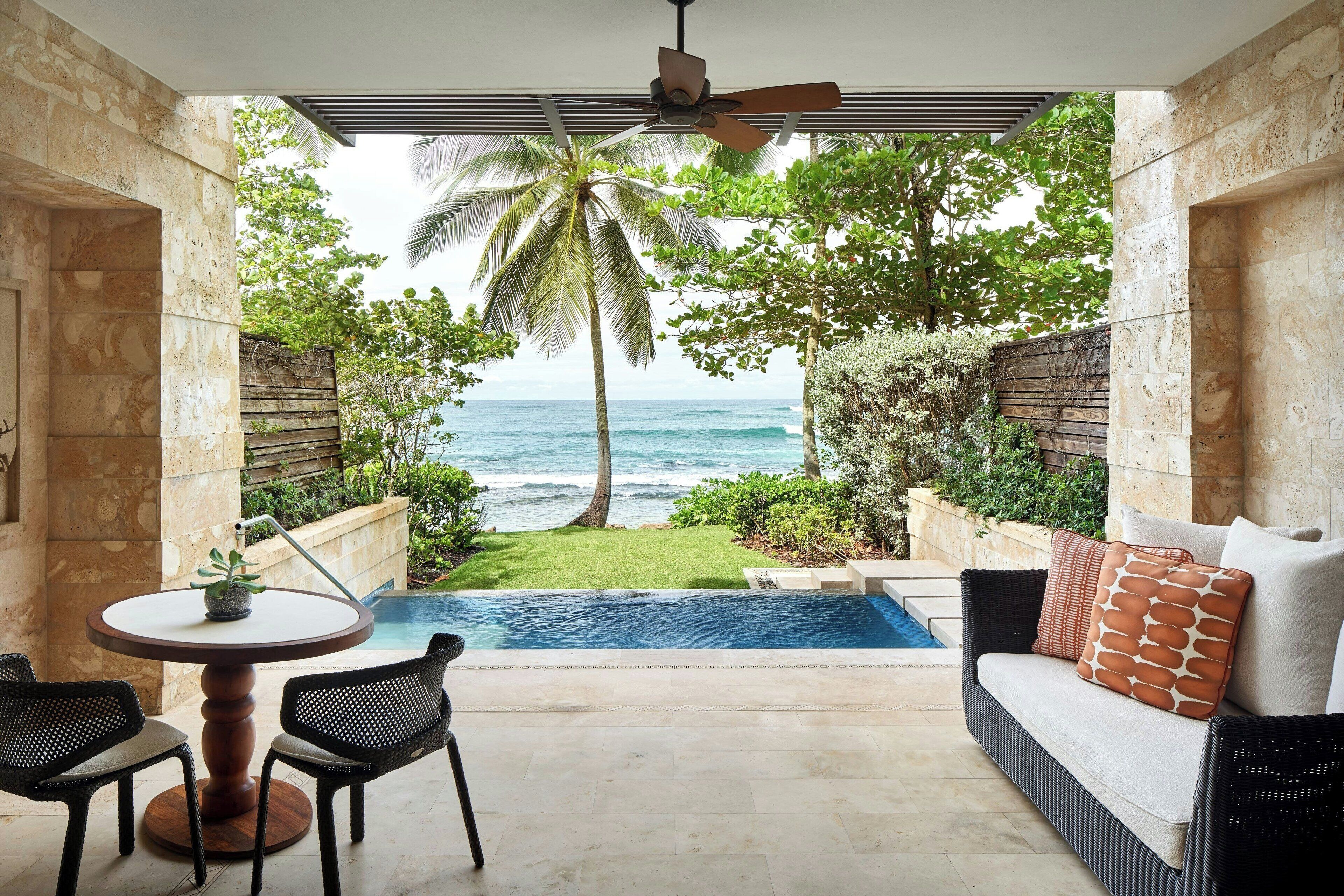 A balcony with a view of the ocean, a pool, and a seating area with a sofa and chairs.