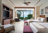 A hotel room with two beds, a TV, a dining area with a table and chairs, and a view of the ocean.
