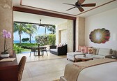 A spacious room with a large bed, a sofa, and a dining area. There is a view of palm trees and the ocean through a glass door.