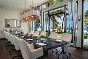 A modern conference room with a large table, white chairs, a hanging light fixture, and a view of the ocean through large windows.