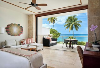 A hotel room with a balcony, a ceiling fan, a large window with a view of the ocean and palm trees, a sofa, a coffee table, and a desk with a plant.