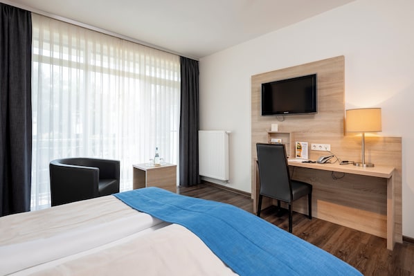 Una habitación de hotel con cama, escritorio, silla, televisor montado en la pared y ventana con cortinas.