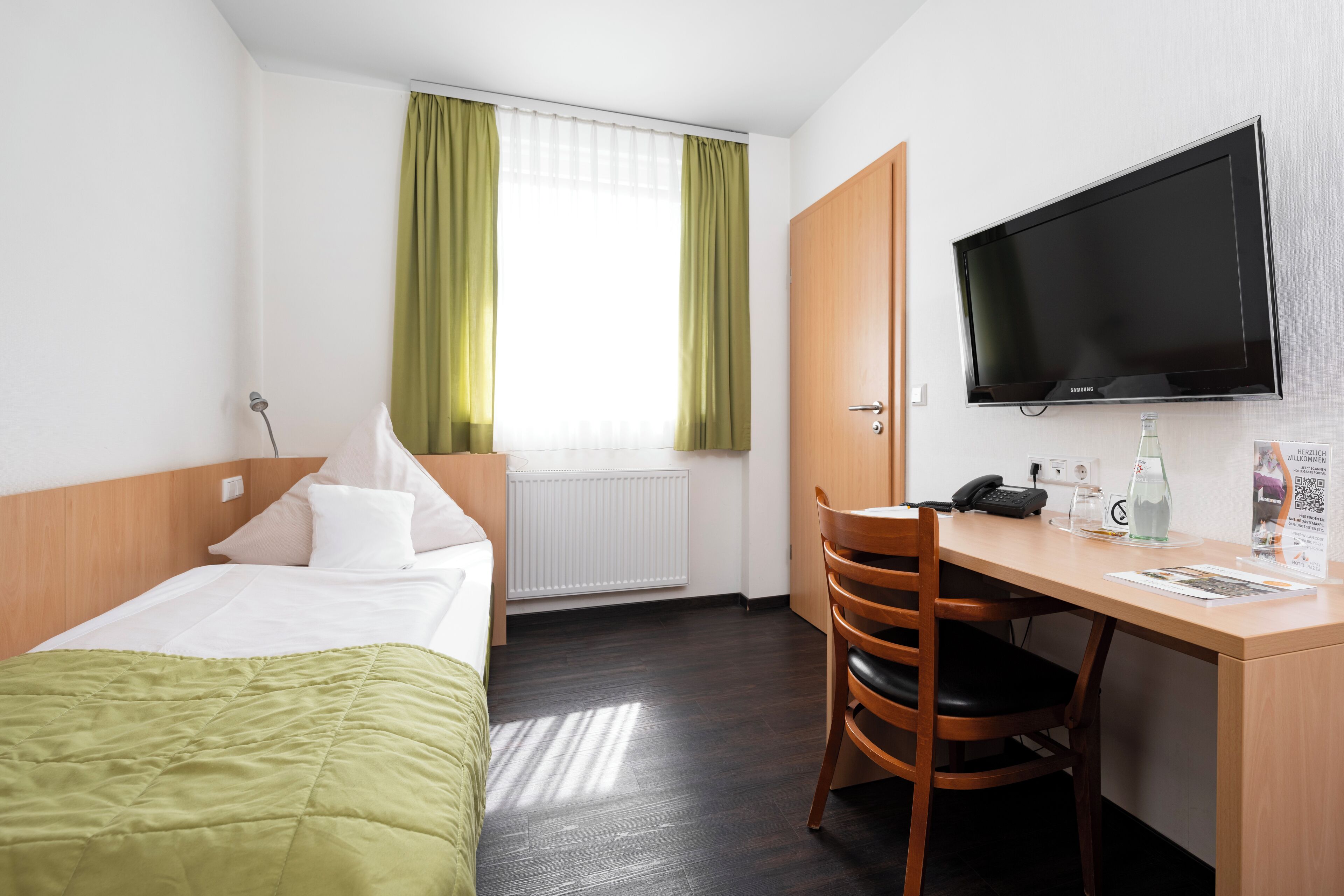 Ett hotellrum med en säng, ett skrivbord med en stol, en tv och ett fönster med gardiner.