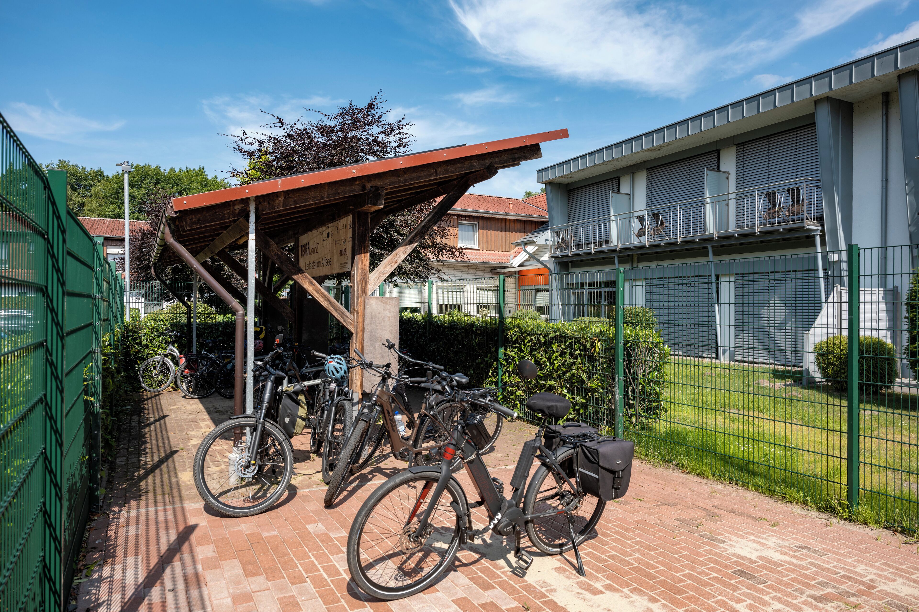 Ett avskärmat område för cykelparkering med cyklar, en gångstig av tegelstenar och en byggnad med ett staket av metall i bakgrunden.