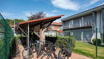 Ett avskärmat område för cykelparkering med cyklar, en gångstig av tegelstenar och en byggnad med ett staket av metall i bakgrunden.