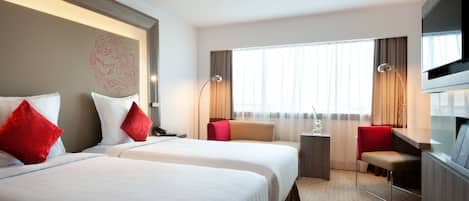 Ett hotellrum med två sängar, ett skrivbord, en stol, en TV och ett fönster med gardiner.