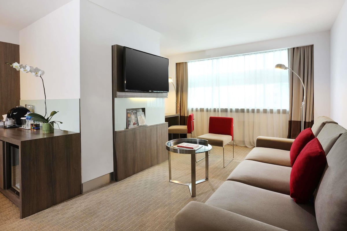 A modern hotel room with a flat-screen TV, a small round table, a sofa with red cushions, a red chair, and a large window with curtains.