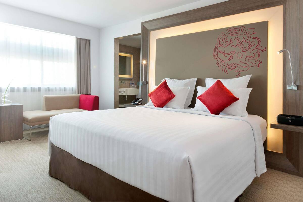 Ein Hotelzimmer mit einem großen Bett, einem Nachttisch, einem Sessel, einem Schreibtisch, einem Spiegel und einem durch eine offene Tür einsehbaren Badezimmer.
