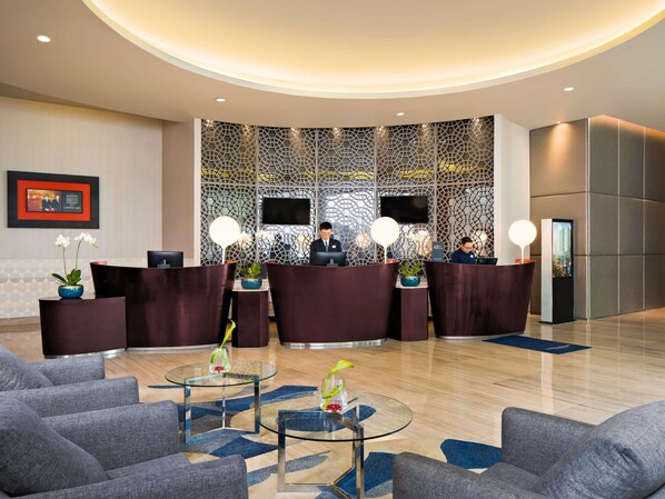 Un hall d’hôtel moderne avec un comptoir d’accueil, deux réceptionnistes et un grand écran de télévision. On y trouve des tables en verre ornées de vases et un sol à motifs.
