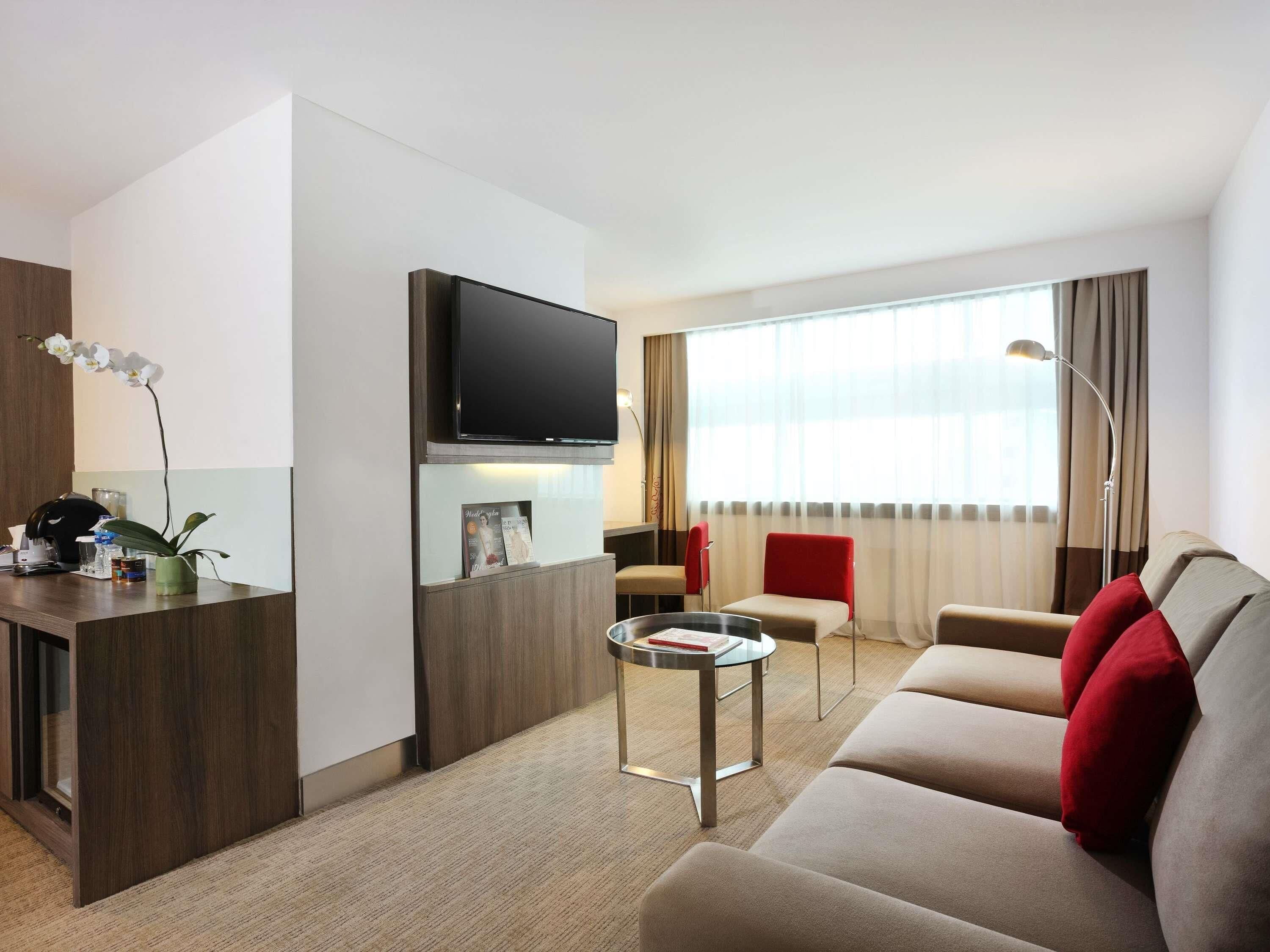 Ein modernes Hotelzimmer mit Flachbildfernseher, einem kleinen runden Tisch, einer Couch mit roten Kissen, einem roten Sessel und einem großen Fenster mit Vorhängen.