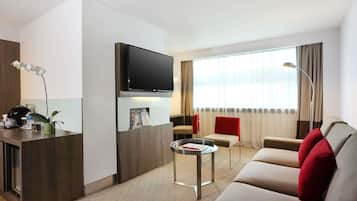 Una habitación de hotel moderna con un televisor de pantalla plana, una pequeña mesa redonda, un sofá con cojines rojos, una silla roja y un ventanal con cortinas.