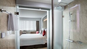 Ein modernes Hotelzimmer mit einer gläsernen Duschkabine, einem ordentlich bezogenen Bett und einem sichtbaren Badezimmerbereich.