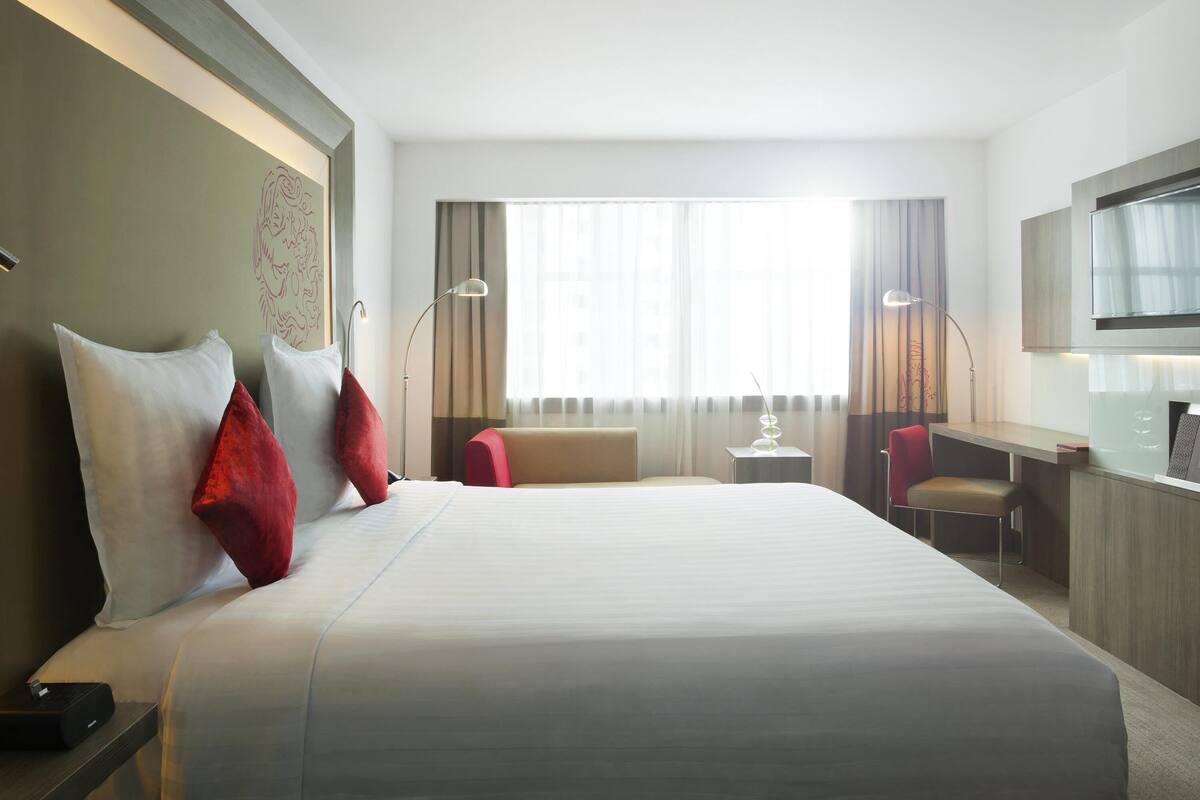 Ein Hotelzimmer mit einem großen Bett, einem Schreibtisch, einem Sessel, einem Fernseher und einem Fenster mit Vorhängen.