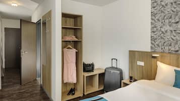 A hotel room with a bed, a wardrobe, a desk, a suitcase, and a wall with a geometric pattern.