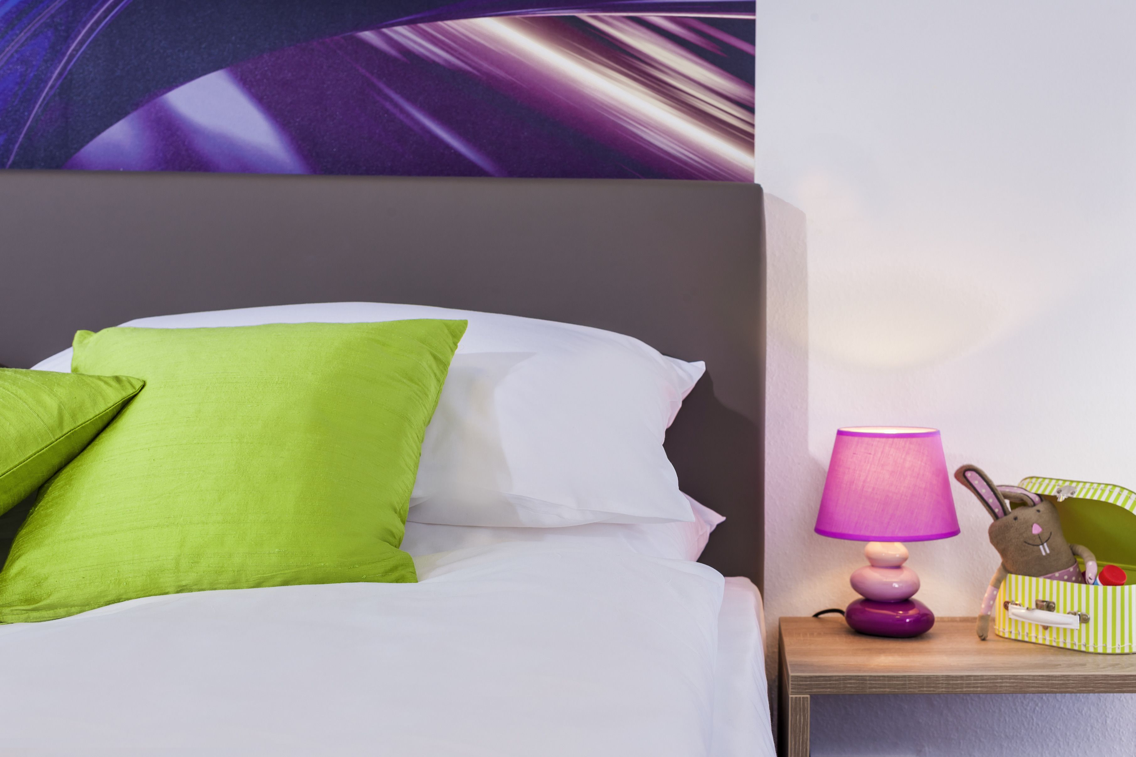 A neatly made bed with green and white pillows, a purple lamp, and a striped storage box with a stuffed toy on a wooden nightstand.