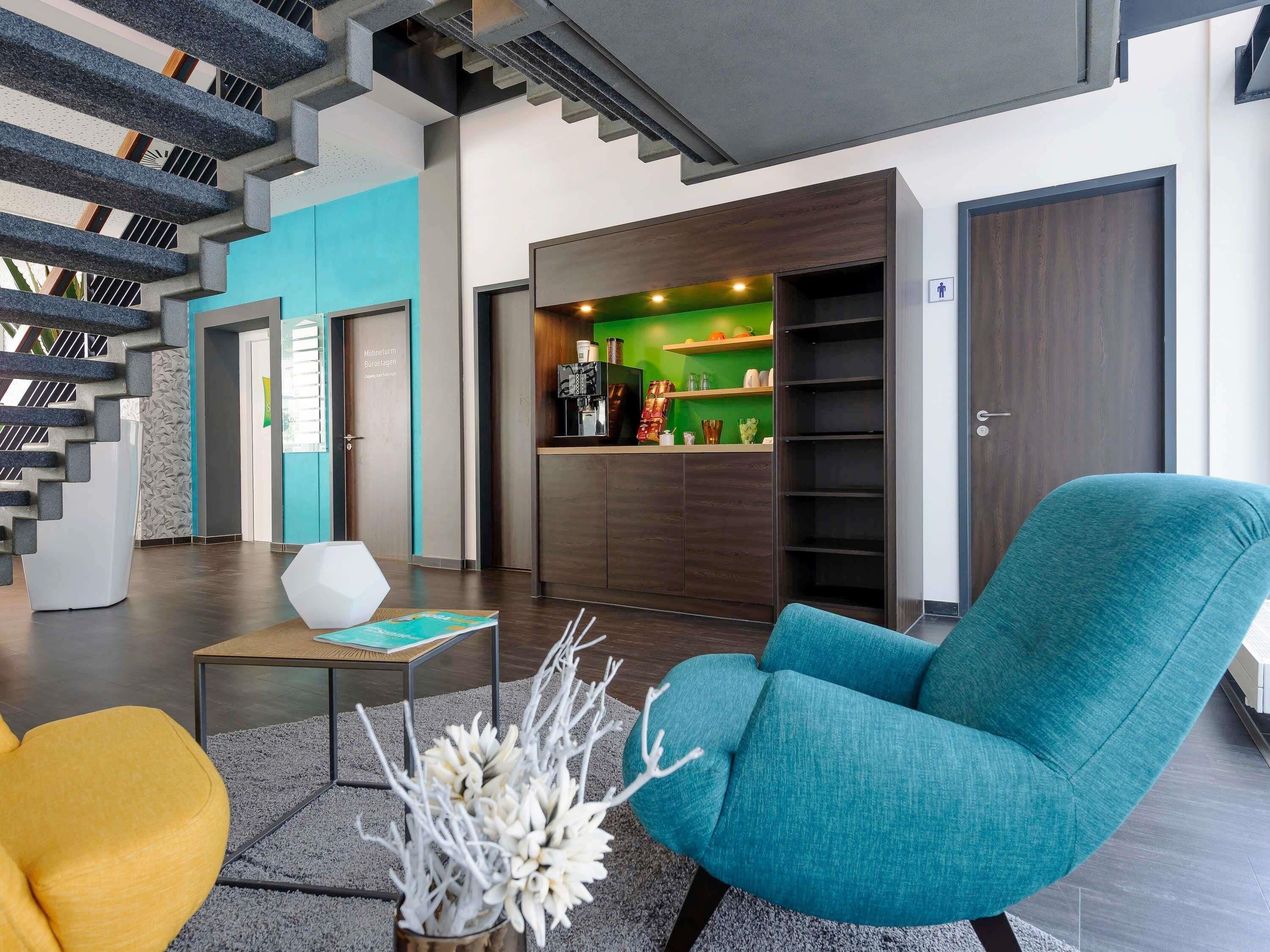 Un vestíbulo moderno con una escalera, un mostrador de recepción, un sillón color turquesa y una cafetera.