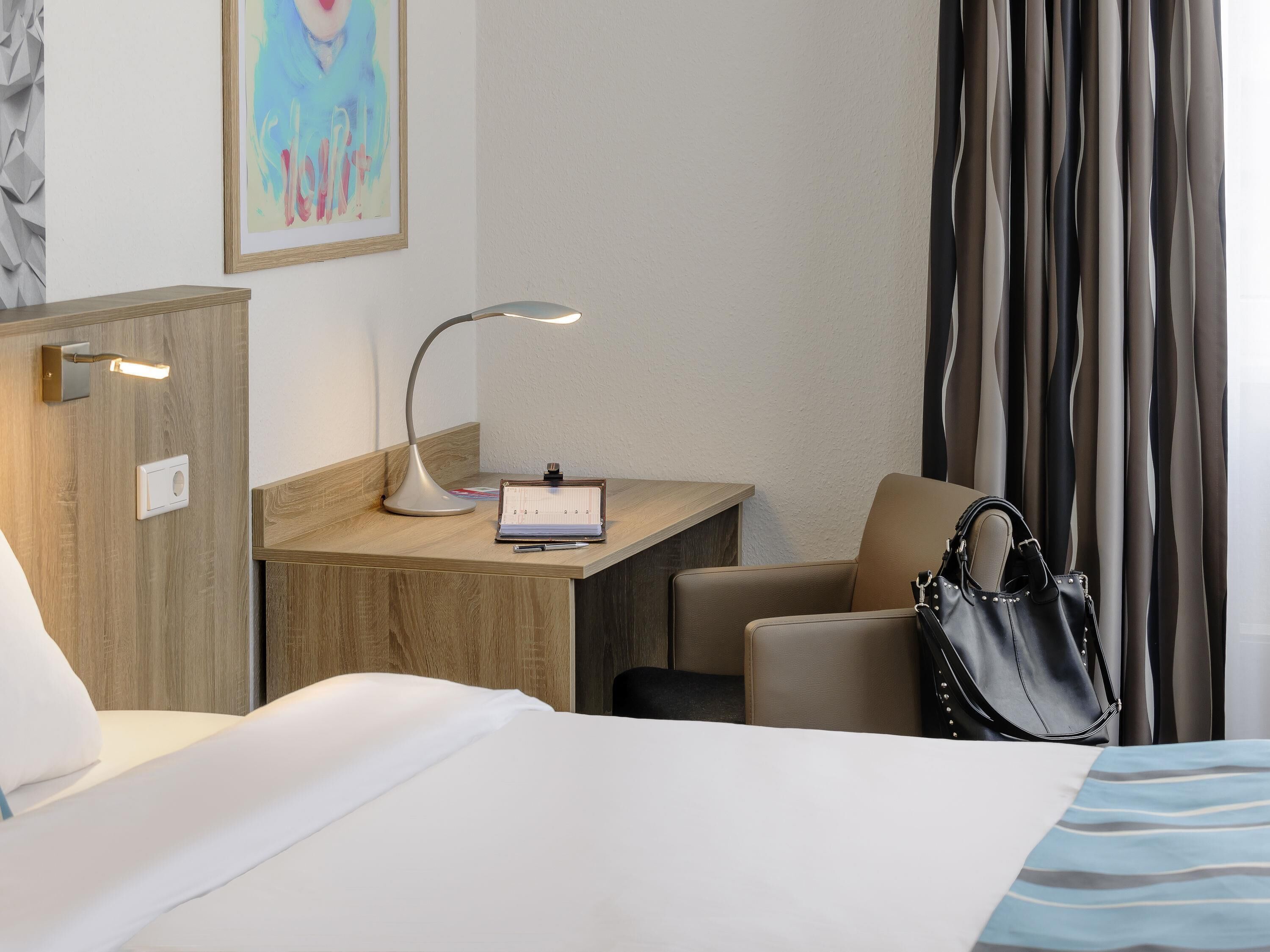 Une chambre d’hôtel avec un lit, un bureau, une chaise, une lampe, un sac à main et un tableau au mur.