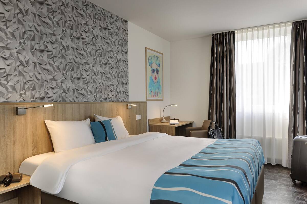 Una camera d'albergo con un letto, comodini, una scrivania, una sedia, una valigia e una finestra con tende.