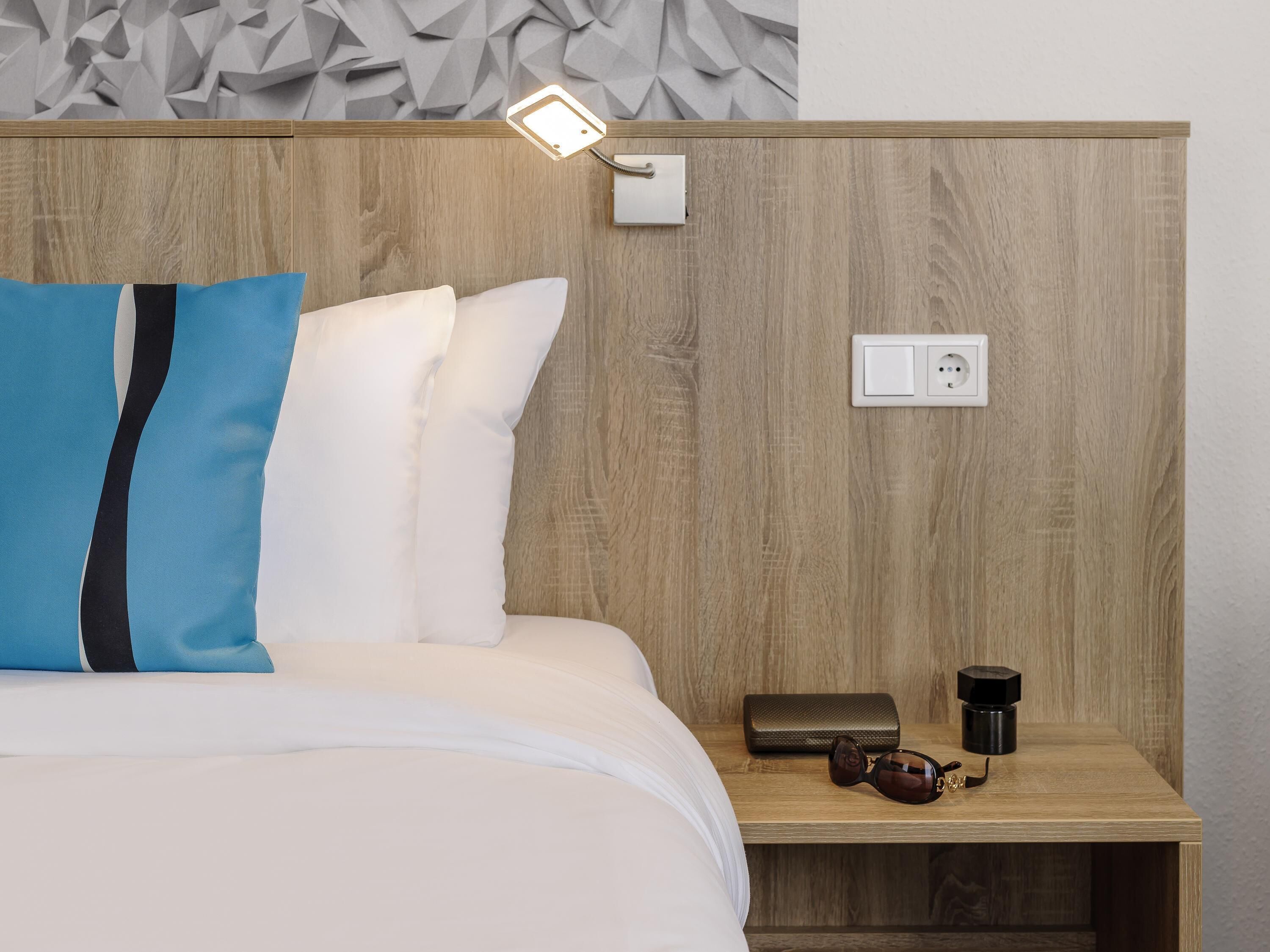 Una cama bien hecha con almohadas blancas y azules, un cabecero de madera, una mesita de noche con una lámpara, un enchufe y un pequeño cofre.