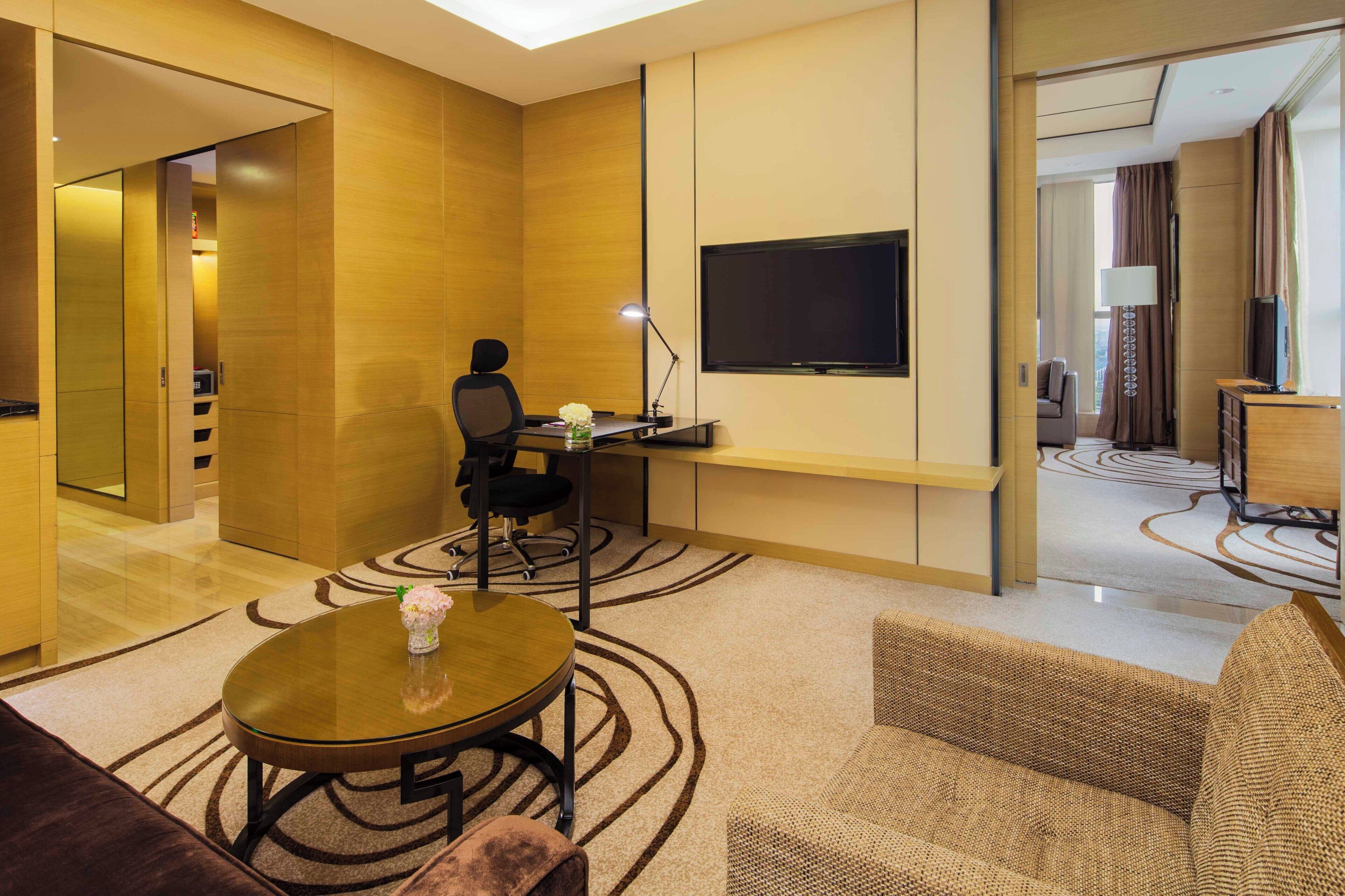 A hotel room with a desk, chair, TV, and a round table with a vase.