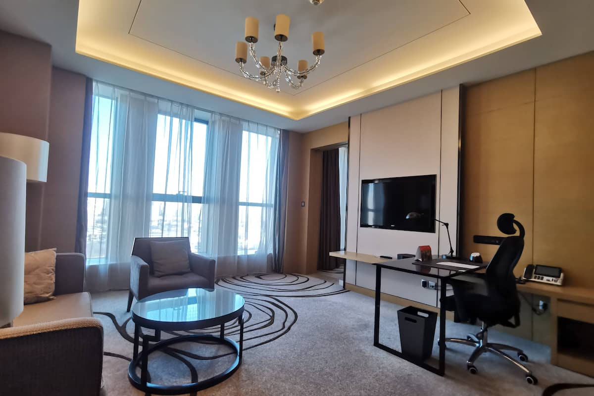 Ein modernes Hotelzimmer mit großem Fernseher, Schreibtisch mit Stuhl, einer Couch und einem runden Couchtisch.