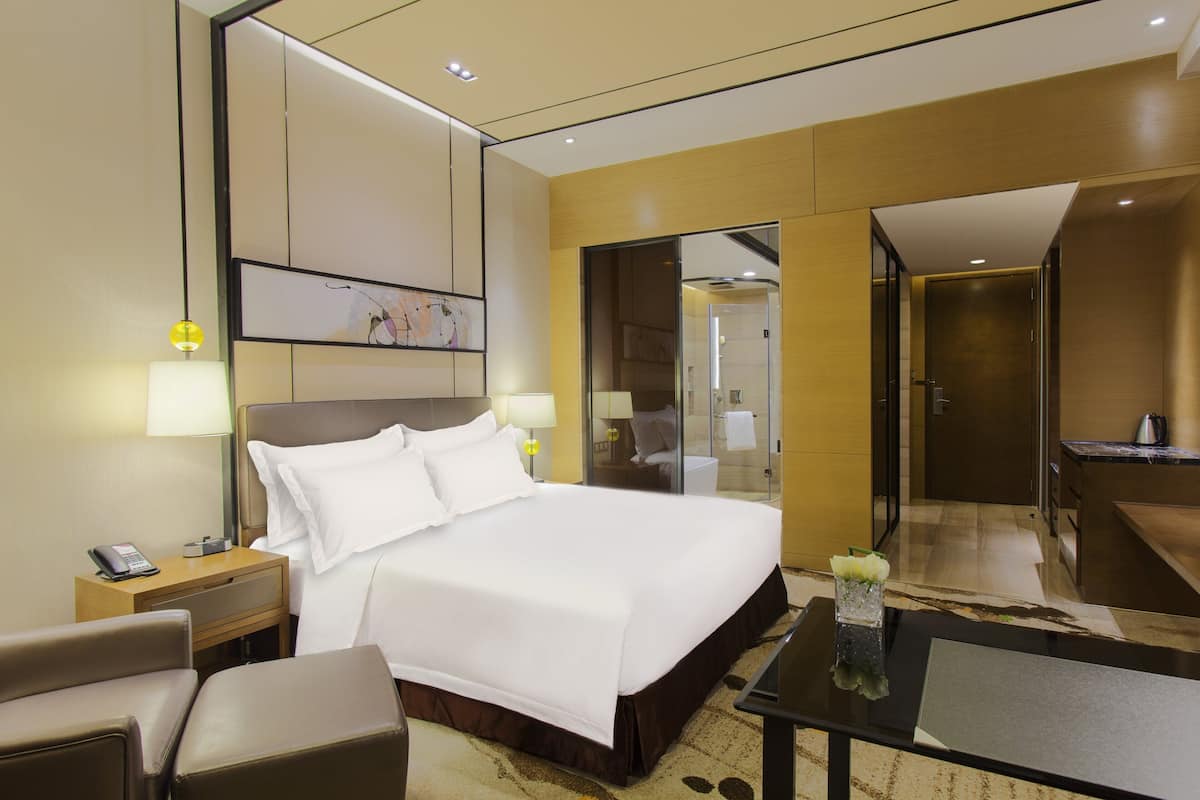 Ein modernes Hotelzimmer mit einem großen Bett, Nachttischen, einem Schreibtisch und einem durch eine Glastür einsehbaren Badezimmer.