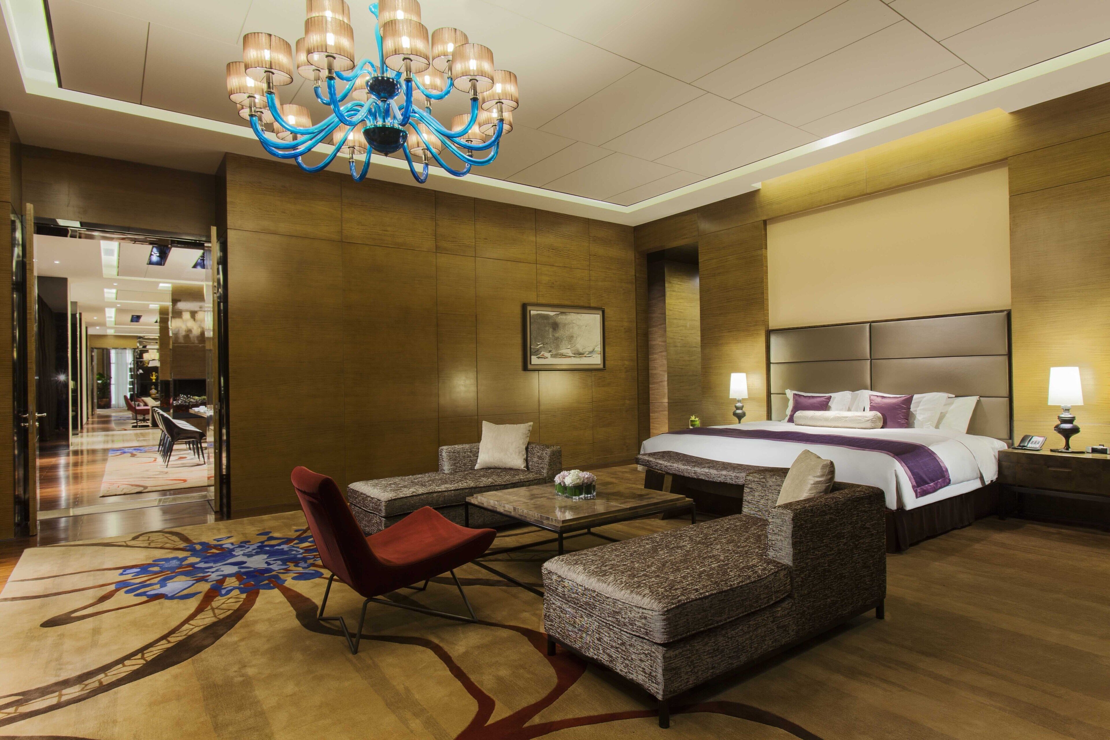 A hotel room with a large bed, a sitting area with a red chair, a coffee table, and a chandelier.