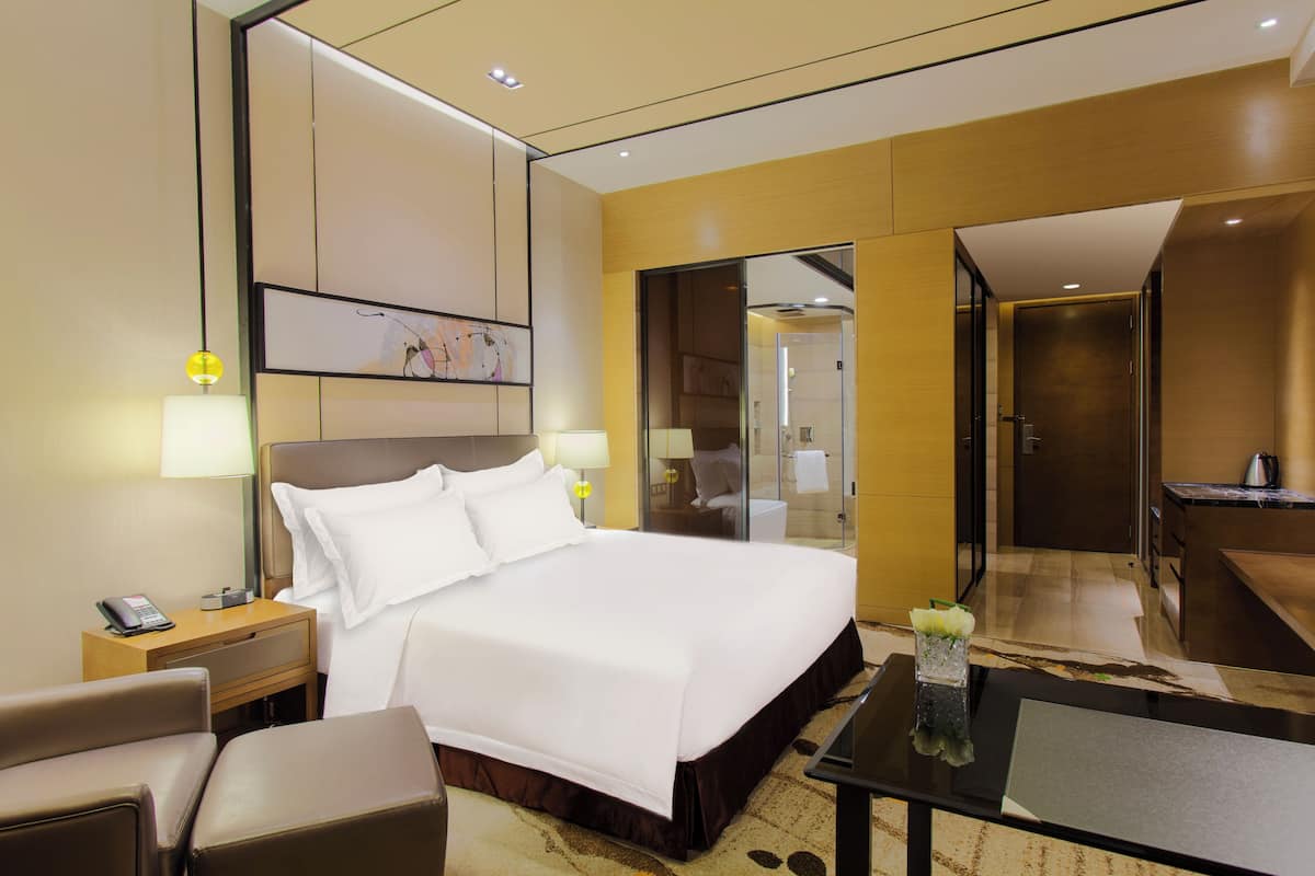 Ein Hotelzimmer mit einem großen Bett, Nachttischen, einem Schreibtisch und einem durch eine Glastür einsehbaren Badezimmer.