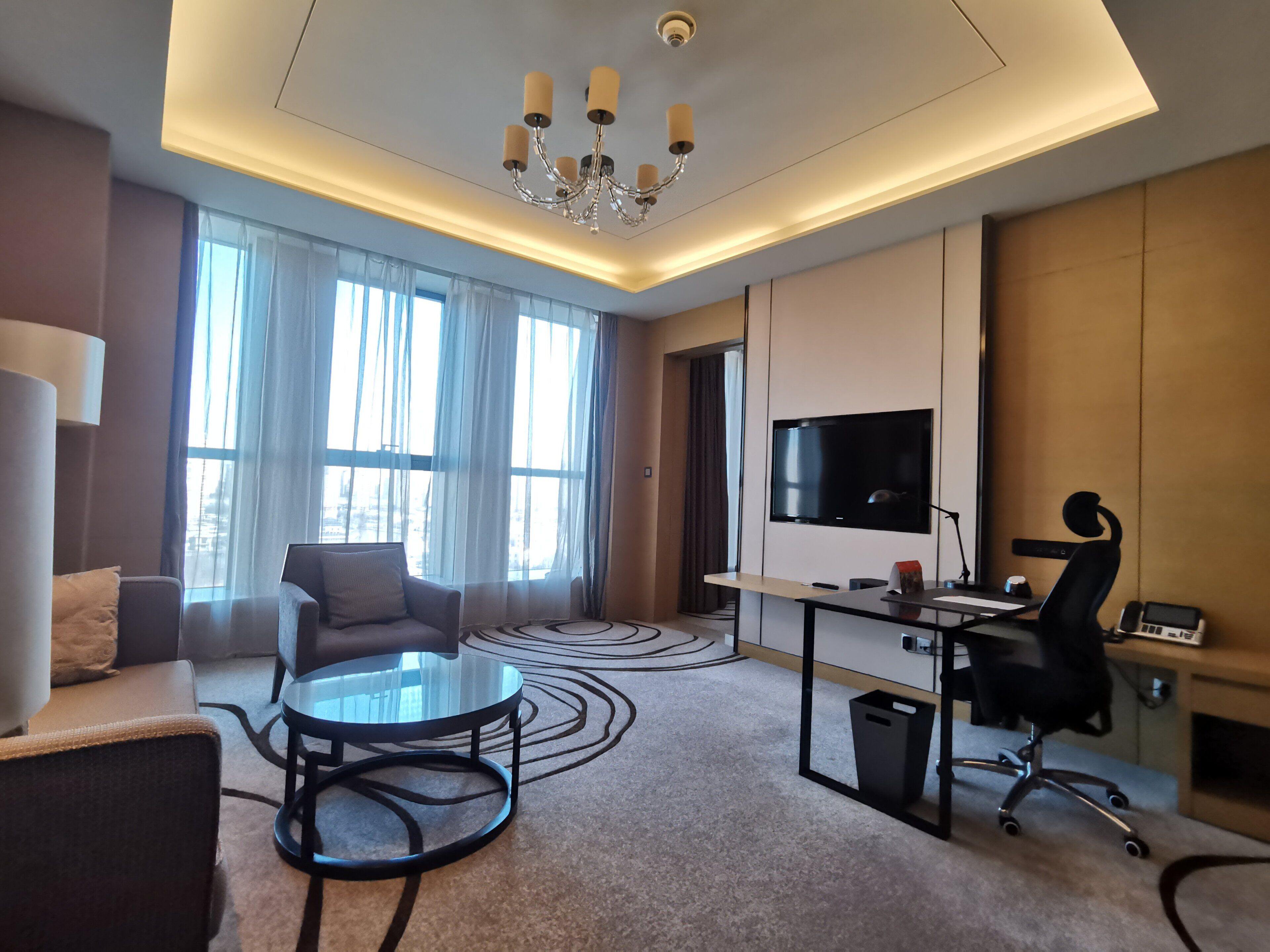 Una camera d'albergo moderna con TV a schermo piatto, una scrivania con sedia, un divano e un lampadario.