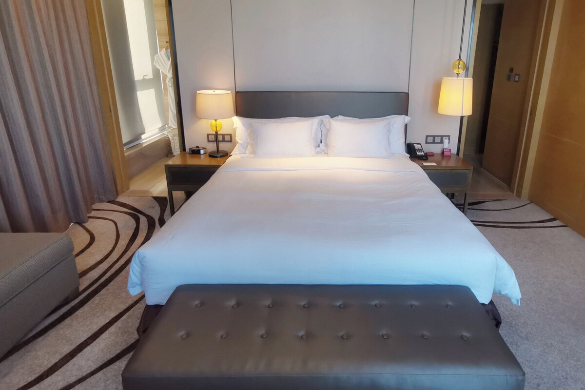 Una habitación de hotel con una cama grande, una mesita de noche con lámpara, una silla y un escritorio pequeño con teléfono y una caja.