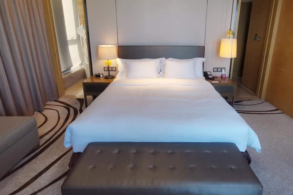 Ein Hotelzimmer mit einem großen Bett, einem Nachttisch mit Lampe, einem Sessel und einem kleinen Schreibtisch mit Telefon und Kasten.